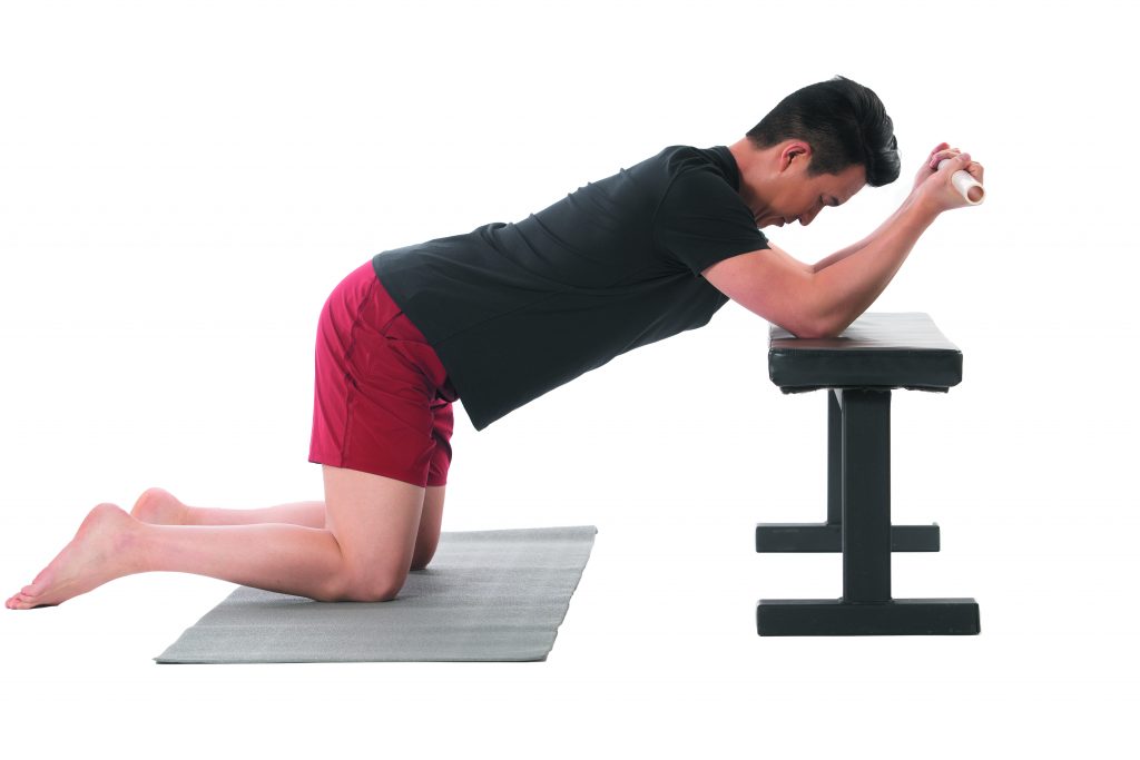 An image is shown of the author kneeling with his elbows on a bench, performing one of the stretches, "T-Spine Extension on a Bench," in his article.