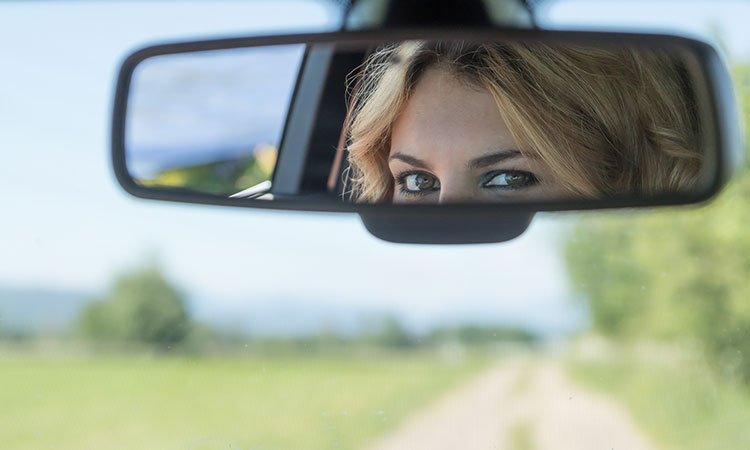 A photo of a person looking into a car's rearview mirror is used to illustrate the idea of the benefit of hindsight.