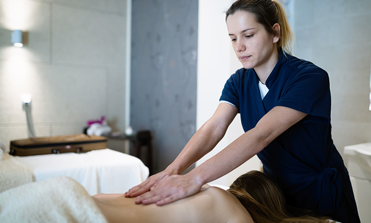 An image of a massage therapist taking a moment to center herself, with both hands on a client's back, is used to illustrate the concept of "holding space" for a massage therapy client.