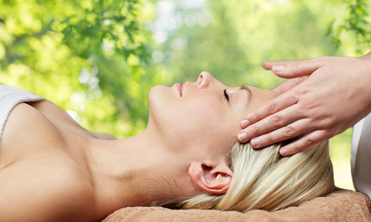 Relaxation massage pictured in a side-view of woman lying with closed eyes and having face or head massaged over green natural background