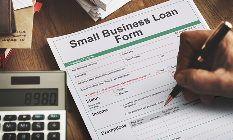 A desk with a calculator and paperwork including a form title Small Business Loan Form that a hand is holding a pen to.