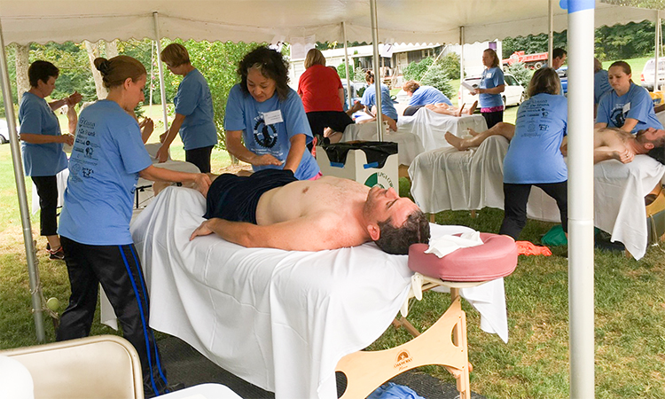10 massage therapists stand while working on clients lying on massage tables, in the white recovery tent on a lawn.